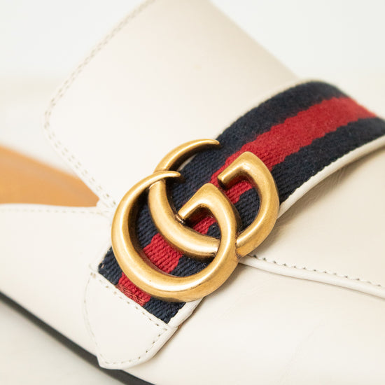 A close-up of a Gucci loafer in off-white leather, featuring the signature red and navy green web stripe and a gold-tone interlocking GG logo buckle.