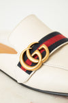A close-up of a Gucci loafer in off-white leather, featuring the signature red and navy green web stripe and a gold-tone interlocking GG logo buckle.
