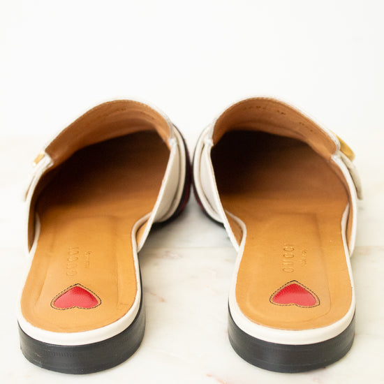 A pair of women's slip-on mule sandals shown from the back, featuring a white leather upper with a small gold buckle, a tan insole with a red heart detail, and a black outsole, set against a light marble background.