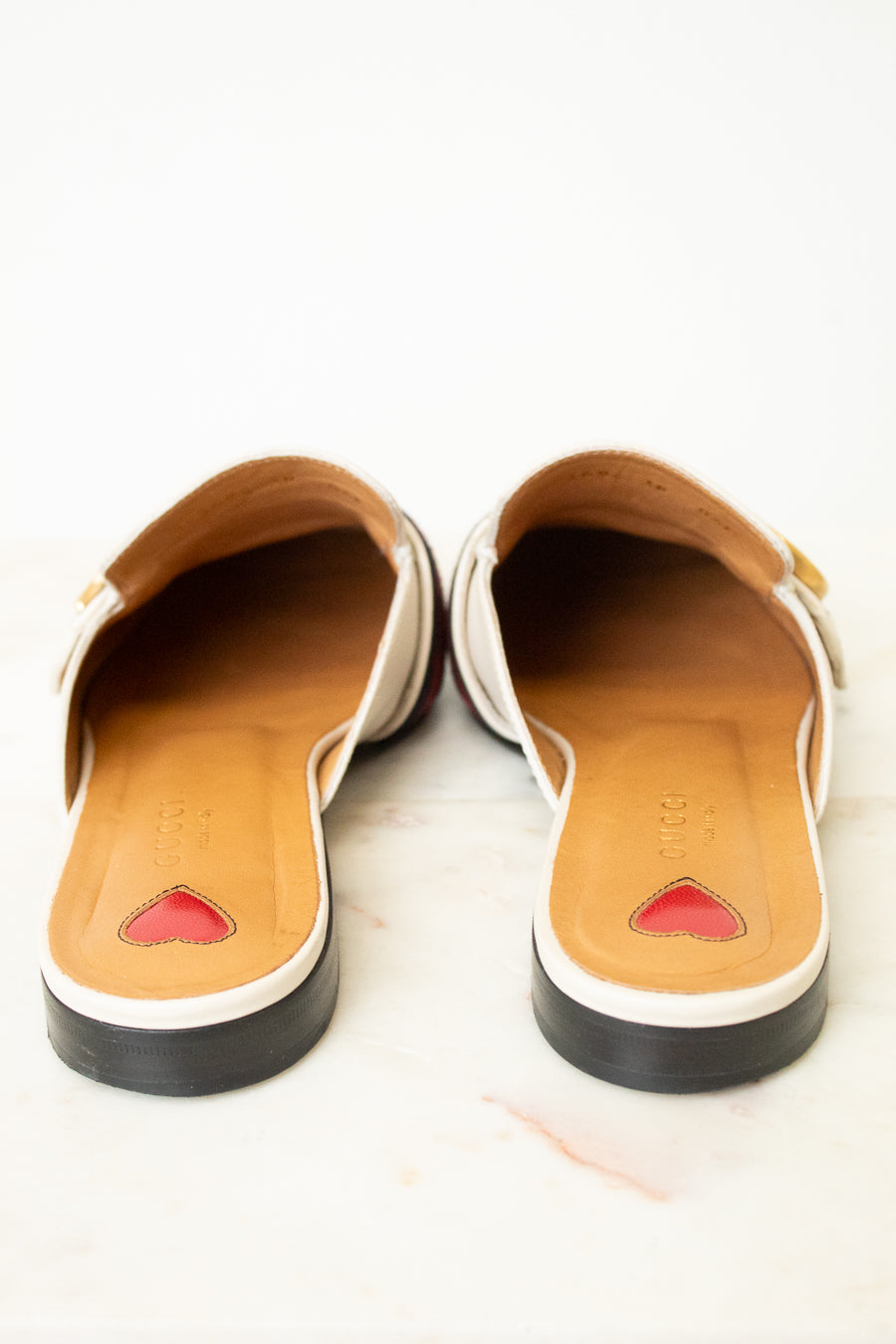 A pair of women's slip-on mule sandals shown from the back, featuring a white leather upper with a small gold buckle, a tan insole with a red heart detail, and a black outsole, set against a light marble background.
