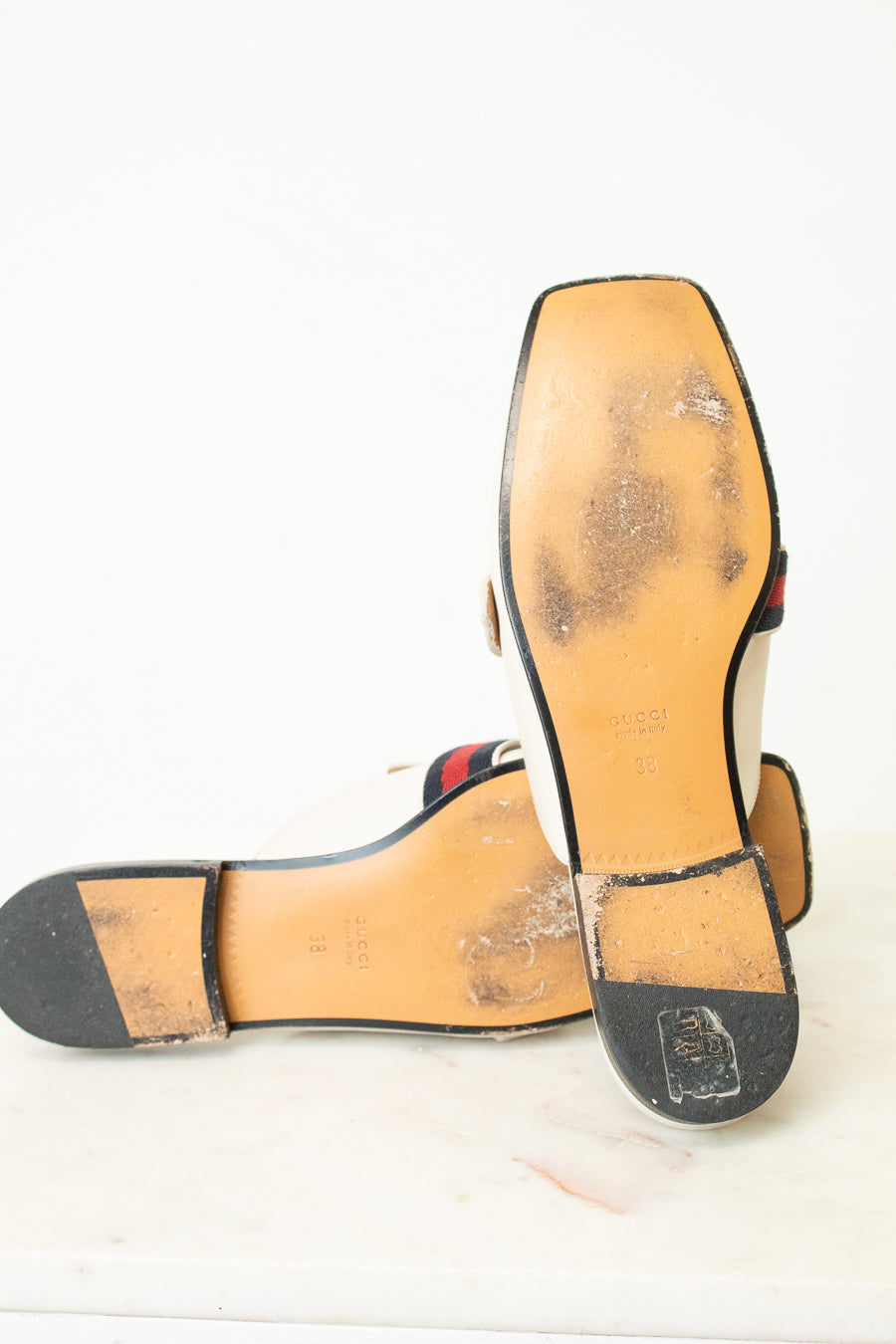 A pair of Gucci flat sandals shown from the sole, displaying the tan leather insoles with heavy scuffing and dirt, black rubber outsoles, and the signature red and green web stripe detail near the heel.