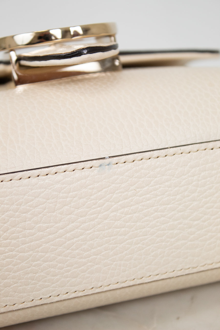 Close-up of a cream-colored pebbled leather handbag with a polished gold-tone metal ring handle and matching stitching.