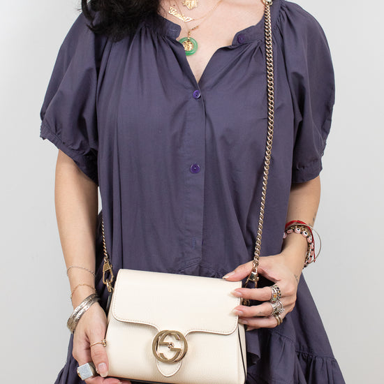 A woman wearing a dark purple puff-sleeve blouse and holding a cream-colored Gucci shoulder bag with a gold-tone horsebit logo clasp and chain strap, set against a plain light gray background.