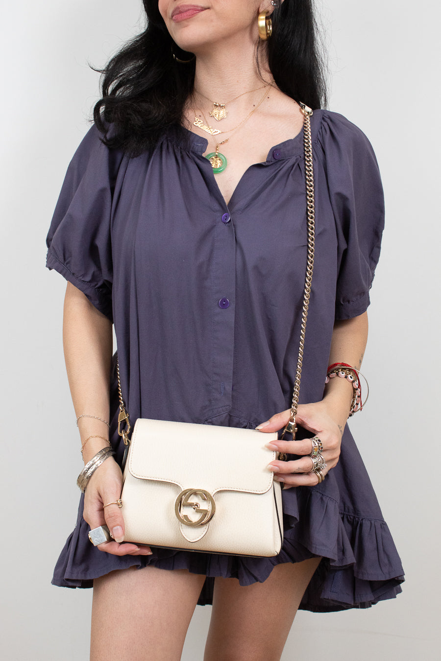 A woman wearing a dark purple puff-sleeve blouse and holding a cream-colored Gucci shoulder bag with a gold-tone horsebit logo clasp and chain strap, set against a plain light gray background.