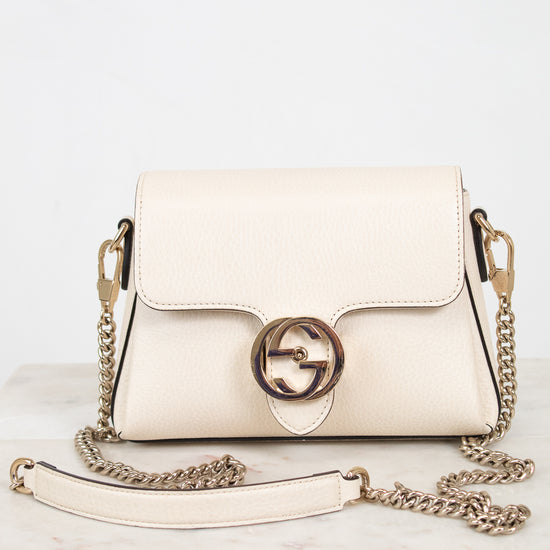 An off-white leather shoulder bag with a gold-tone interlocking G logo clasp and a matching chain strap, displayed on a light marble surface against a white background.