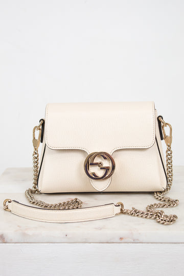 An off-white leather shoulder bag with a gold-tone interlocking G logo clasp and a matching chain strap, displayed on a light marble surface against a white background.