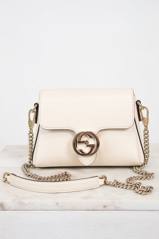 An off-white leather shoulder bag with a gold-tone interlocking G logo clasp and a matching chain strap, displayed on a light marble surface against a white background.