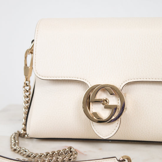 An off-white leather shoulder bag with a gold-tone interlocking G logo clasp and a matching chain strap, resting on a light marble surface.