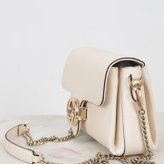 A cream-colored leather shoulder bag with gold-tone hardware and a chain strap, resting on a light marble surface against a white background.