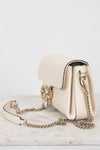 A cream-colored leather shoulder bag with gold-tone hardware and a chain strap, resting on a light marble surface against a white background.