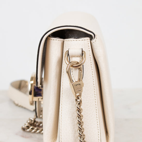 A cream-colored leather shoulder bag with gold-tone hardware and a gold chain strap, shown from a side profile view on a light marble surface against a white background.