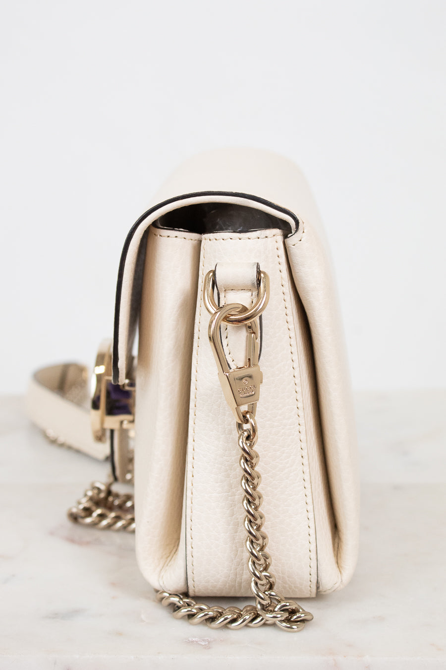 A cream-colored leather shoulder bag with gold-tone hardware and a gold chain strap, shown from a side profile view on a light marble surface against a white background.