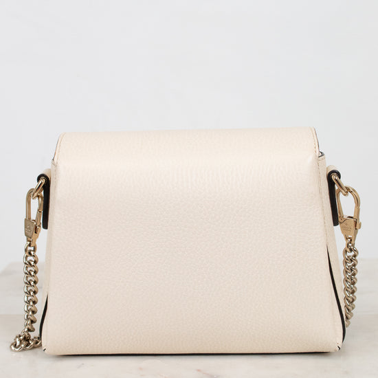 A cream-colored pebbled leather crossbody bag with a gold-tone chain strap, displayed on a white marble surface against a white background.