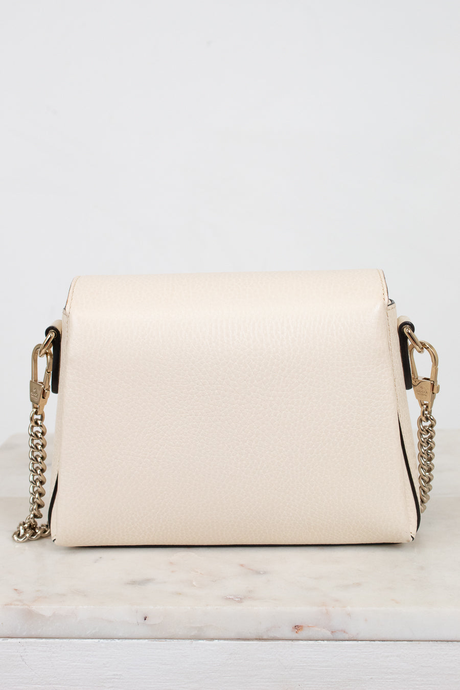 A cream-colored pebbled leather crossbody bag with a gold-tone chain strap, displayed on a white marble surface against a white background.