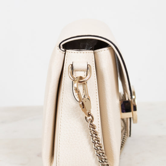 A cream-colored pebbled leather handbag shown from a side profile view, featuring a gold-tone chain strap and a black leather trim accent, resting on a light marble surface against a white background.