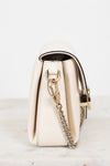 A cream-colored pebbled leather handbag shown from a side profile view, featuring a gold-tone chain strap and a black leather trim accent, resting on a light marble surface against a white background.