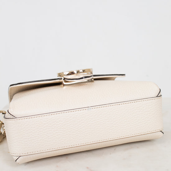 A small cream-colored pebbled leather handbag with a gold-tone metal clasp, resting on a white marble surface against a white background.