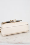 A small cream-colored pebbled leather handbag with a gold-tone metal clasp, resting on a white marble surface against a white background.