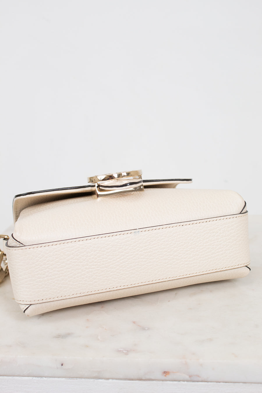 A small cream-colored pebbled leather handbag with a gold-tone metal clasp, resting on a white marble surface against a white background.