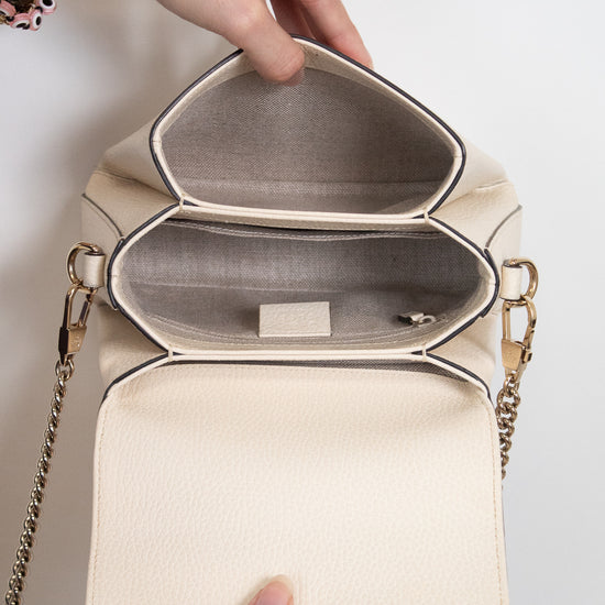 An off-white pebbled leather handbag with gold-tone chain straps, shown open to reveal a structured interior with two compartments. A person's hands are holding the bag open against a plain white background.
