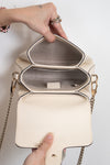 An off-white pebbled leather handbag with gold-tone chain straps, shown open to reveal a structured interior with two compartments. A person's hands are holding the bag open against a plain white background.