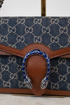 Close-up of a denim handbag with brown leather details and blue and silver buckle on a white background