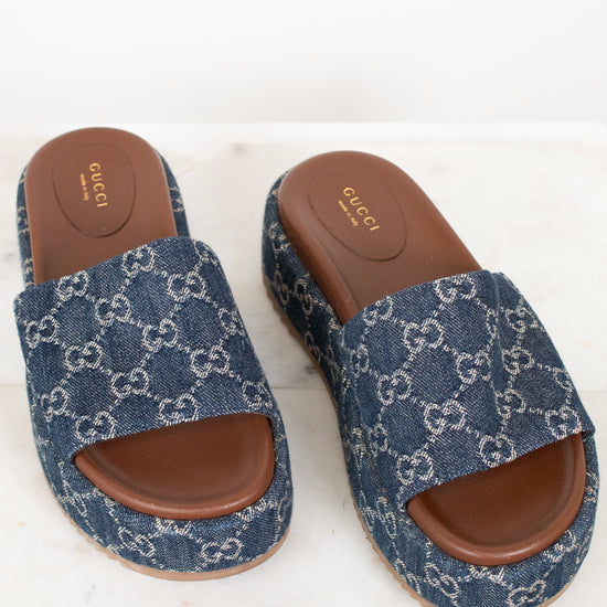A pair of Gucci slide sandals featuring a blue and white GG monogram canvas upper with a brown leather footbed and sole, displayed on a light marble surface.