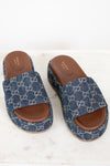 A pair of Gucci slide sandals featuring a blue and white GG monogram canvas upper with a brown leather footbed and sole, displayed on a light marble surface.