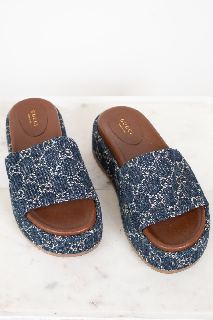 A pair of Gucci slide sandals featuring a blue and white GG monogram canvas upper with a brown leather footbed and sole, displayed on a light marble surface.