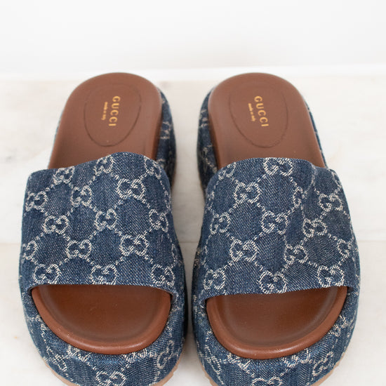 A pair of Gucci slide sandals featuring a blue and white GG monogram denim upper with brown leather soles, displayed on a light marble surface against a white background.
