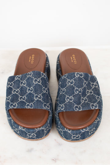 A pair of Gucci slide sandals featuring a blue and white GG monogram denim upper with brown leather soles, displayed on a light marble surface against a white background.