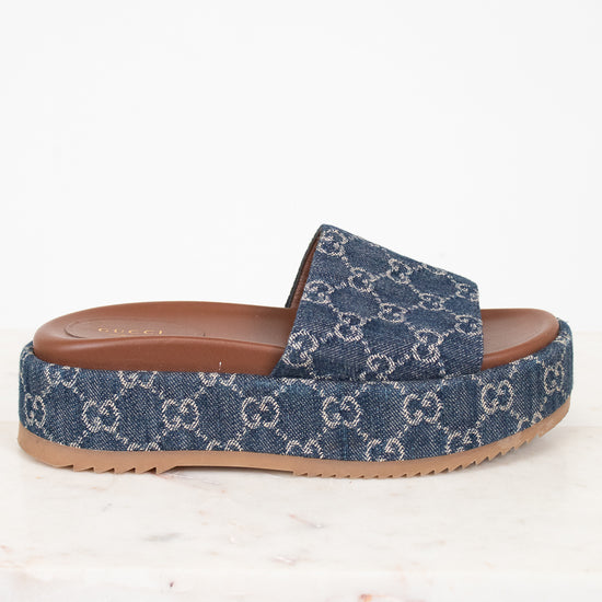A single Gucci platform slide sandal in blue GG Supreme denim with a brown leather insole and tan rubber sole, shown from the side on a white marble surface against a white background.