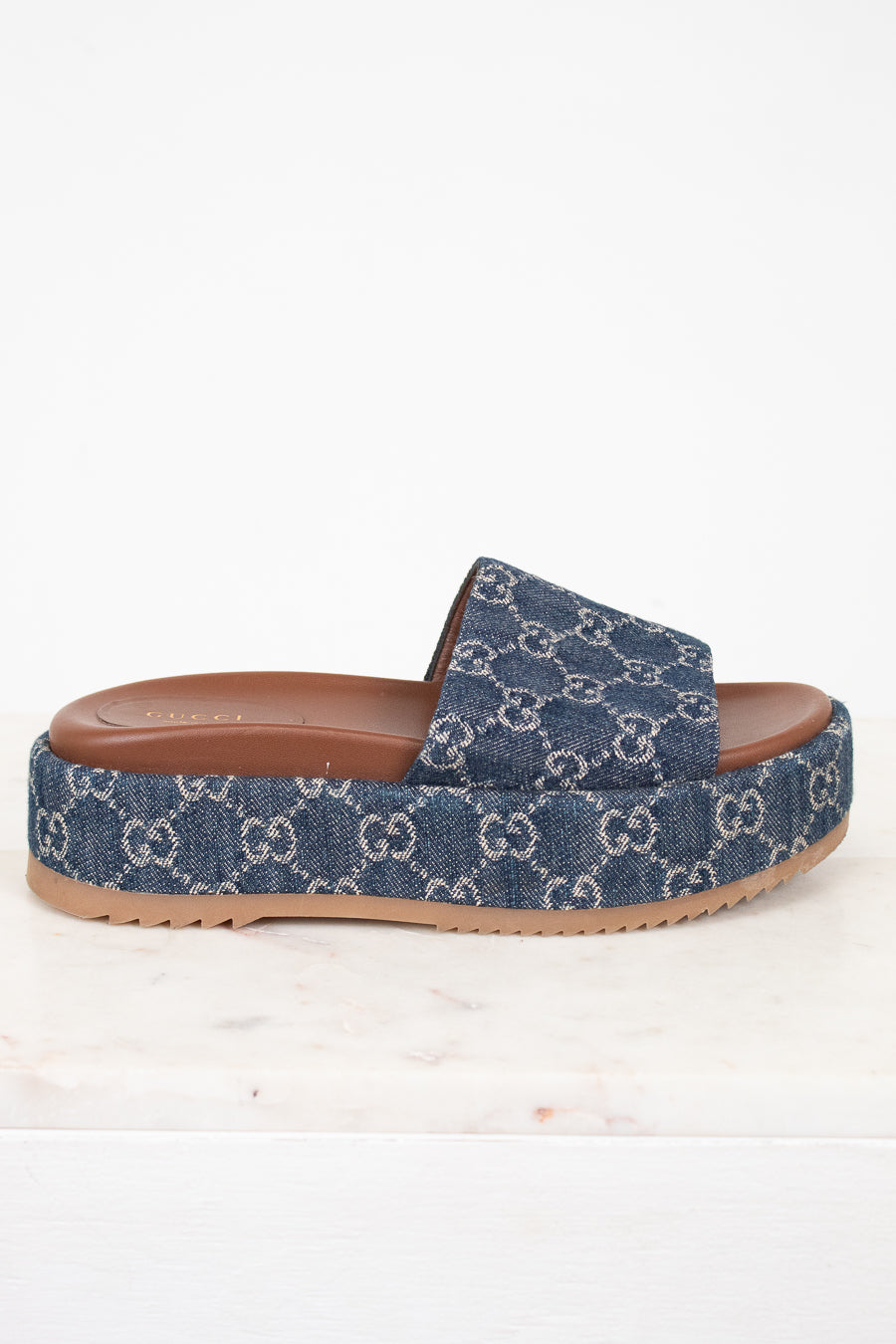 A single Gucci platform slide sandal in blue GG Supreme denim with a brown leather insole and tan rubber sole, shown from the side on a white marble surface against a white background.