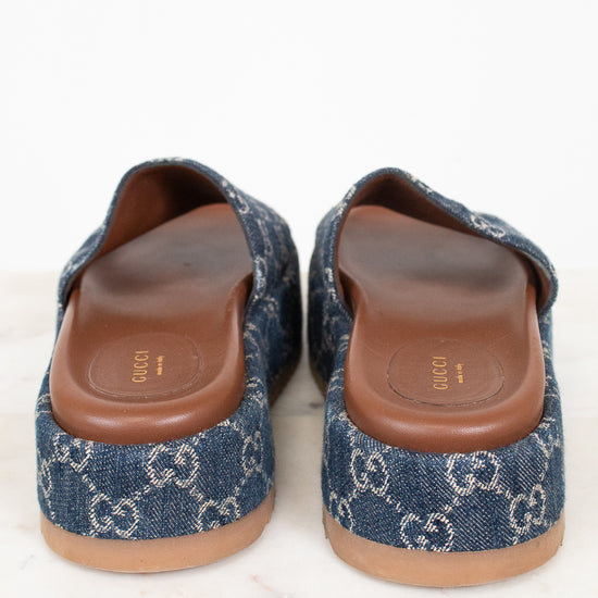 A pair of platform Gucci slip-on mule sandals made of blue denim with the brand's signature GG monogram pattern, featuring brown leather insoles and tan soles, photographed from the back on a white marble surface.