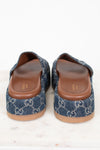 A pair of platform Gucci slip-on mule sandals made of blue denim with the brand's signature GG monogram pattern, featuring brown leather insoles and tan soles, photographed from the back on a white marble surface.