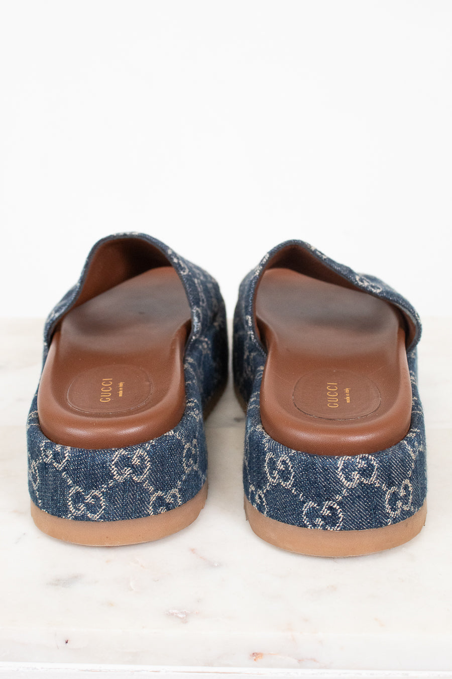 A pair of platform Gucci slip-on mule sandals made of blue denim with the brand's signature GG monogram pattern, featuring brown leather insoles and tan soles, photographed from the back on a white marble surface.
