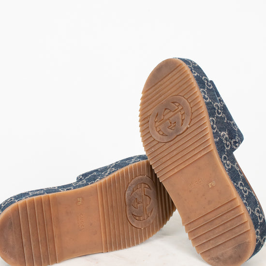 A pair of Gucci platform slip-on shoes shown from the bottom, featuring a navy blue monogram denim upper and a brown rubber sole with an embossed Gucci logo.