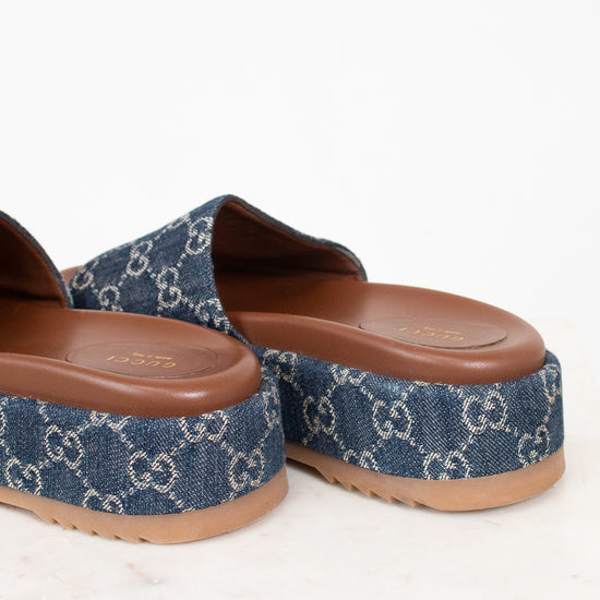 A pair of Gucci platform slide sandals in blue denim with the signature GG monogram pattern, featuring a brown footbed and tan sole, shown on a light marble surface against a white background.