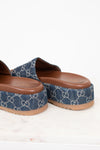 A pair of Gucci platform slide sandals in blue denim with the signature GG monogram pattern, featuring a brown footbed and tan sole, shown on a light marble surface against a white background.