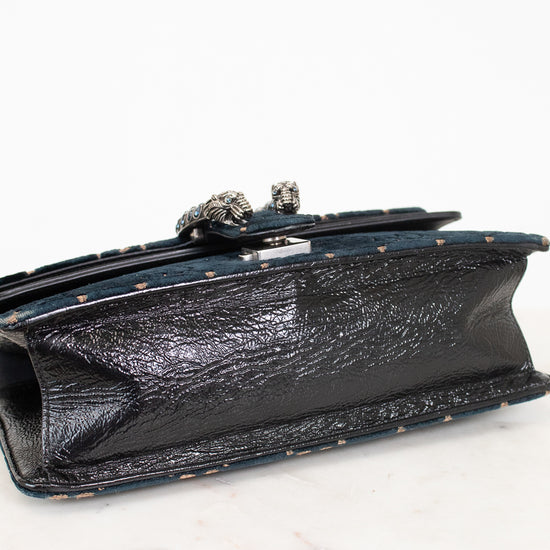 A close-up of the bottom of a shoulder bag showing a black textured leather panel with a silver-tone decorative metal clasp, resting on a white marble surface against a white background.