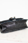 A close-up of the bottom of a shoulder bag showing a black textured leather panel with a silver-tone decorative metal clasp, resting on a white marble surface against a white background.