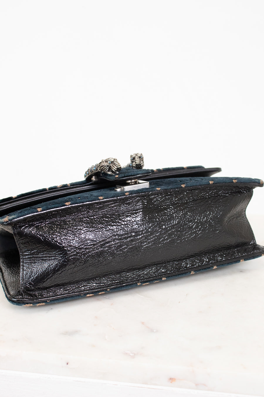 A close-up of the bottom of a shoulder bag showing a black textured leather panel with a silver-tone decorative metal clasp, resting on a white marble surface against a white background.