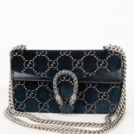A dark teal velvet shoulder bag with a gold-tone GG monogram pattern, a silver chain strap and a silver-tone chain strap with a rhinestone-embellished Gucci dionysus clasp, shown on a white marble surface against a white background.