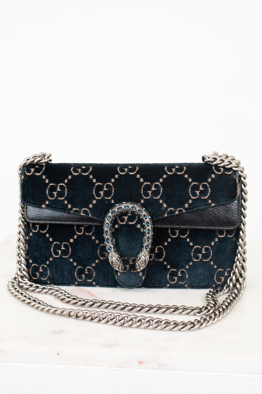 A dark teal velvet shoulder bag with a gold-tone GG monogram pattern, a silver chain strap and a silver-tone chain strap with a rhinestone-embellished Gucci dionysus clasp, shown on a white marble surface against a white background.