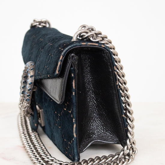A dark teal blue velvet and black leather shoulder bag with a silver-tone chain strap, shown from a side angle on a light marble surface against a white background.