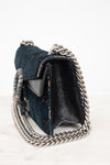 A dark teal blue velvet and black leather shoulder bag with a silver-tone chain strap, shown from a side angle on a light marble surface against a white background.