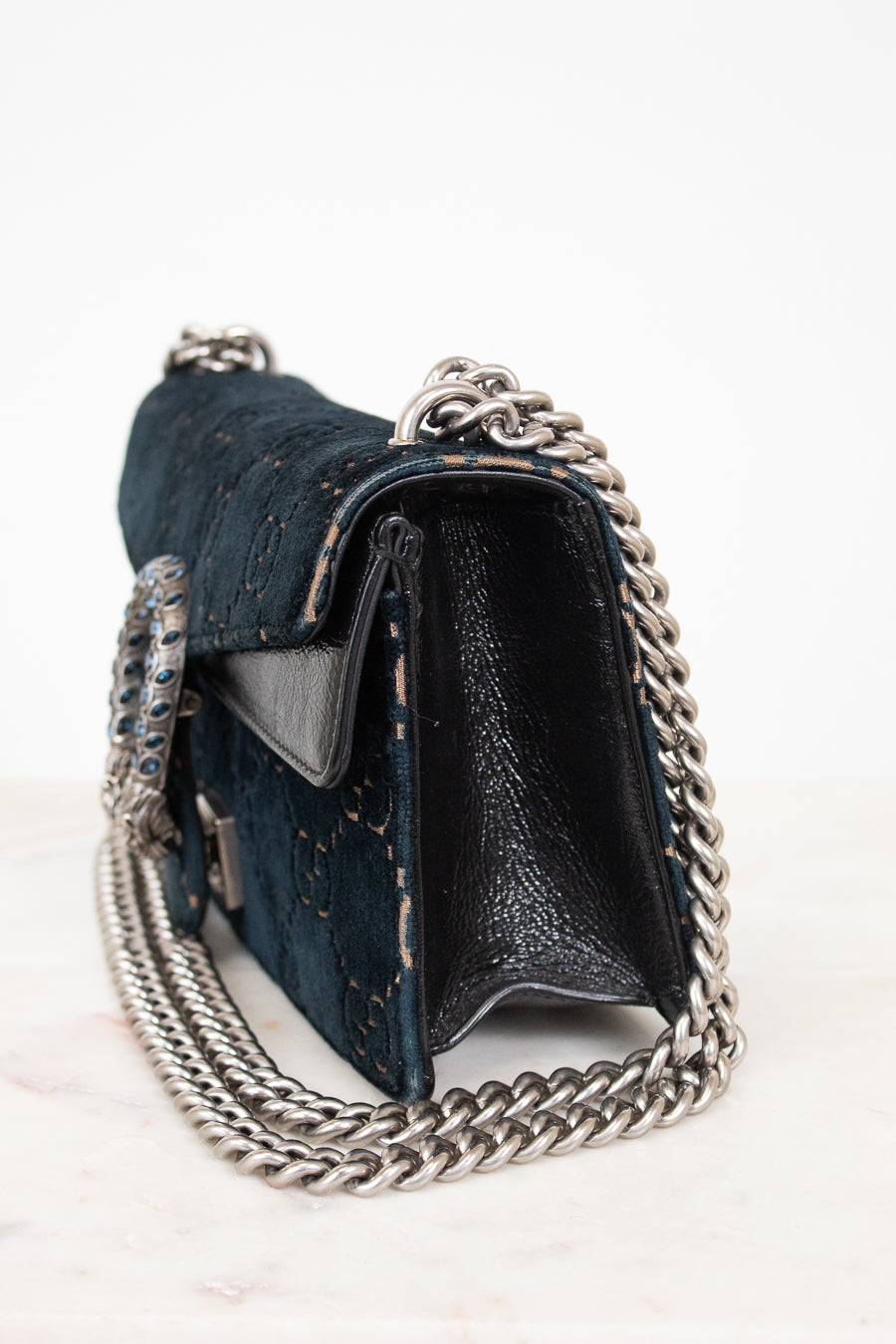 A dark teal blue velvet and black leather shoulder bag with a silver-tone chain strap, shown from a side angle on a light marble surface against a white background.