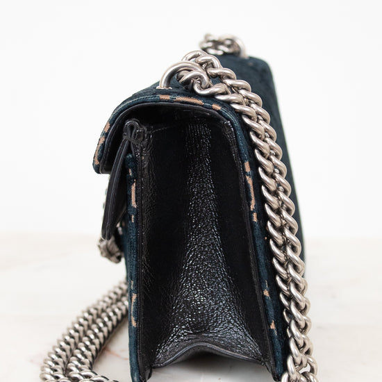 A close-up side view of a black leather handbag with a chunky silver chain strap, resting on a light marble surface against a white background.