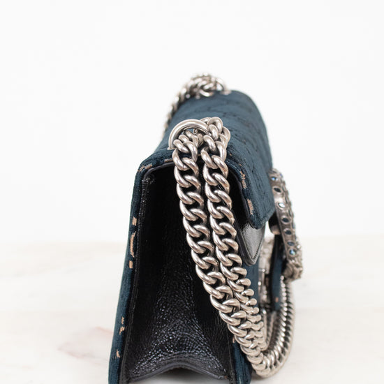 A close-up side view of a dark teal textured leather handbag with a chunky silver chain strap, resting on a white marble surface against a white background.