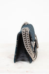 A close-up side view of a dark teal textured leather handbag with a chunky silver chain strap, resting on a white marble surface against a white background.
